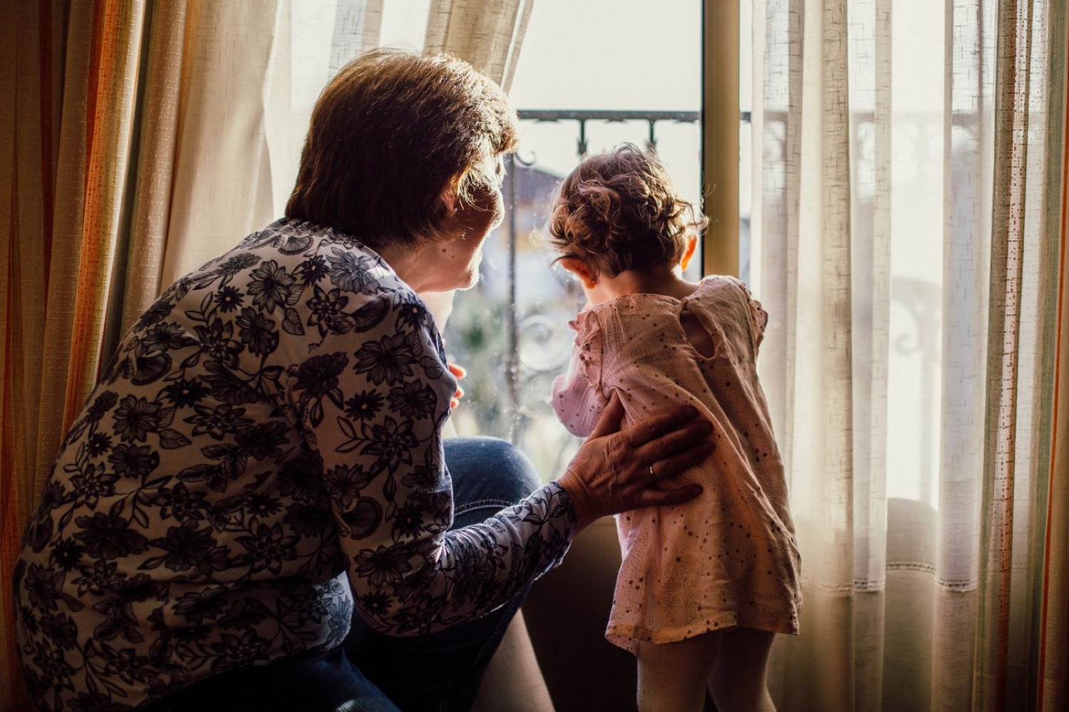 An older woman crouches beside a small child at a sunlit window, one hand resting on the child's back.